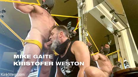 Mike Gaite stands naked, bound in bright yellow rope. A chain is attached to a leather collar around his neck which holds a metal hook, set deep in his ass. Kristofer Weston sits back stroking his cock through his shorts...